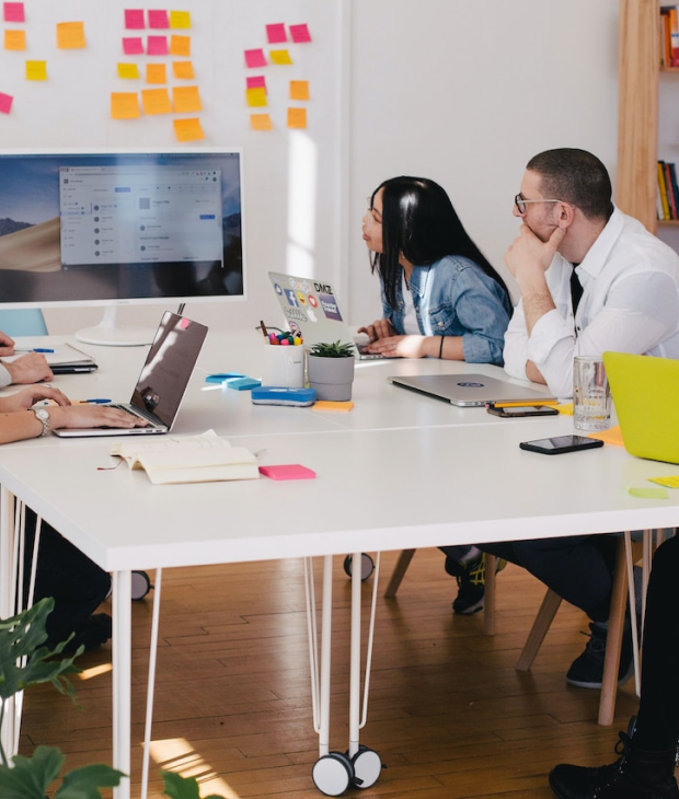 A group of workers at a table, leaning in to see what one coworker is displaying on the monitor for them.