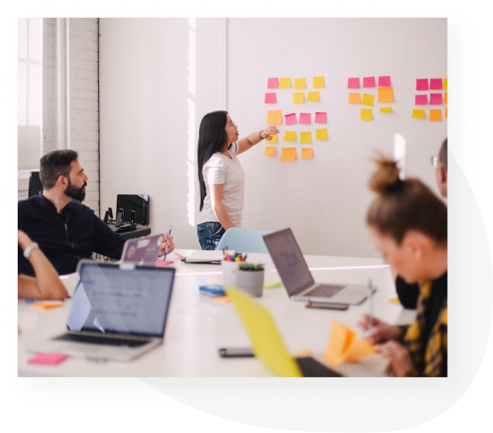 In an office, a woman stands before a group and goes over the multicolored Post-It notes stuck on the wall as they plan the future of their business.