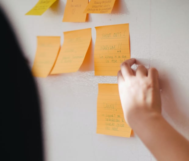 A hand reaching to touch or move one of several Post-It notes displayed on an office wall.